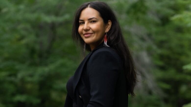 A medium profile shot of a long-haired person facing the camera, smiling, wearing beaded earrings.