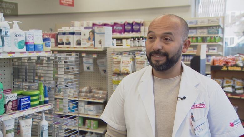 A man in a white coat stands in a pharmacy aisle.