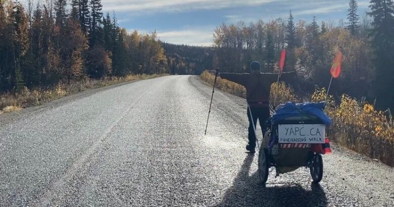 A man is walking down the highway with his back to the camera. His arms are stretched out on either side of his body. He is pulling a cart with a sign on the back that says Y A P C DOT C A fundraising walk.  It's a beautiful fall morning.