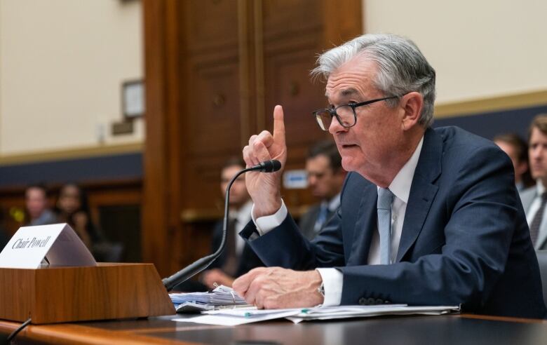 A man with white hair and glasses, and wearing a blue suit, points his right index finger in the air as he sits at a desk and speaks into a microphone.