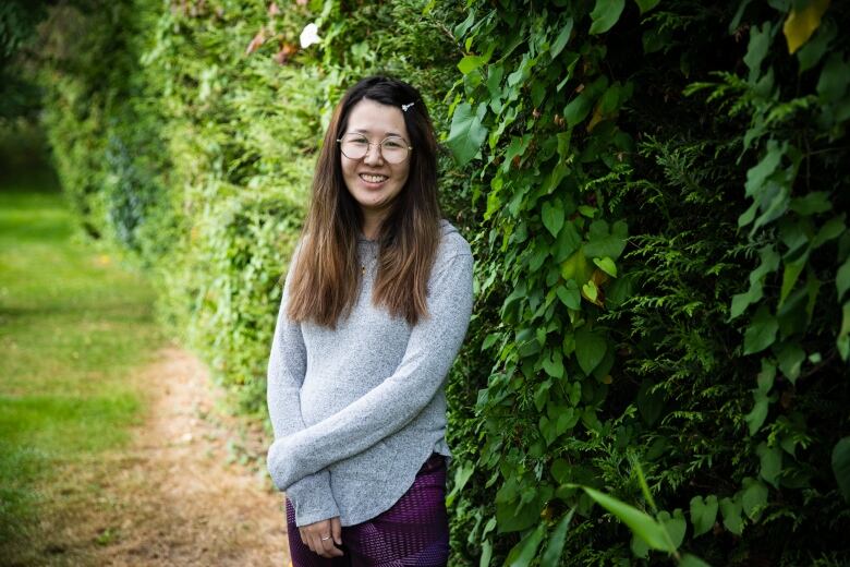 A young East Asian woman with glasses and long hair stands in front of a green hedge.