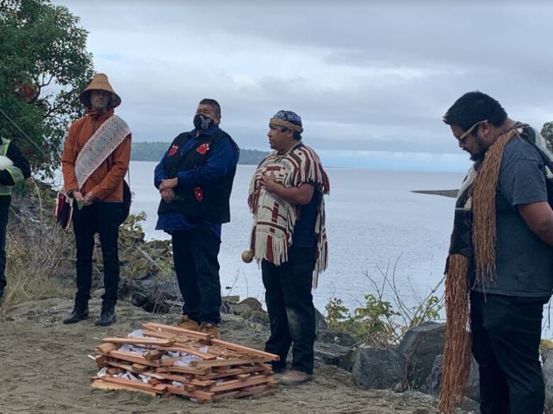 A number of people stand around a woodstack near a riverbank.