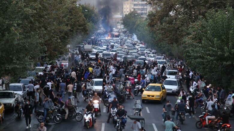 A crowd, including people, scooters, motorcycles and vehicles, blocks a road.