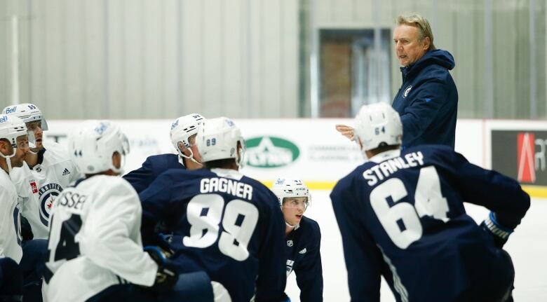 A group of Winnipeg Jets kneel on the ice while listening to new coach Rick Bowness, who is standing, talking and gesturing.