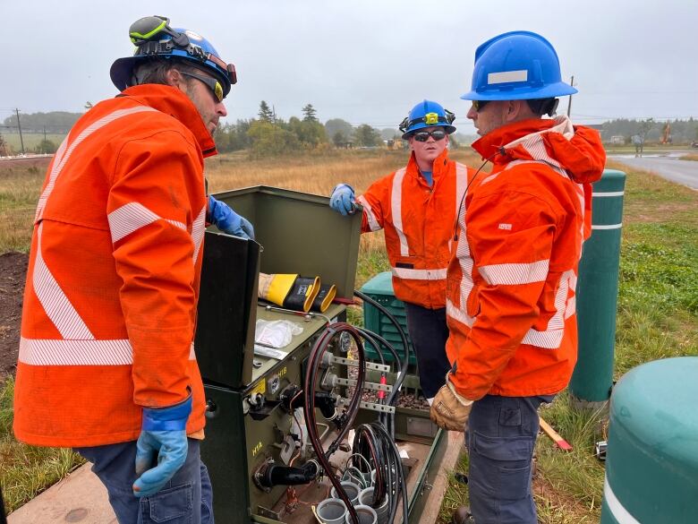 Three men in orange safety jackets confer around a piece of electrical equipment. 