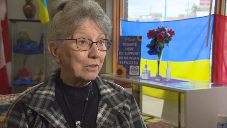 A woman with grey hair and glasses stands inside a room. A Ukrainian flag is hung up in the background.