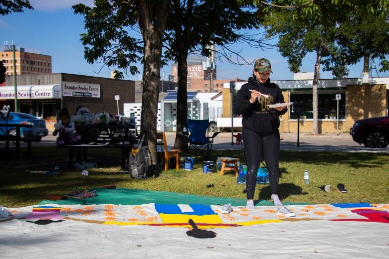 A woman stands holding a paintbrush for a big mural on the ground.