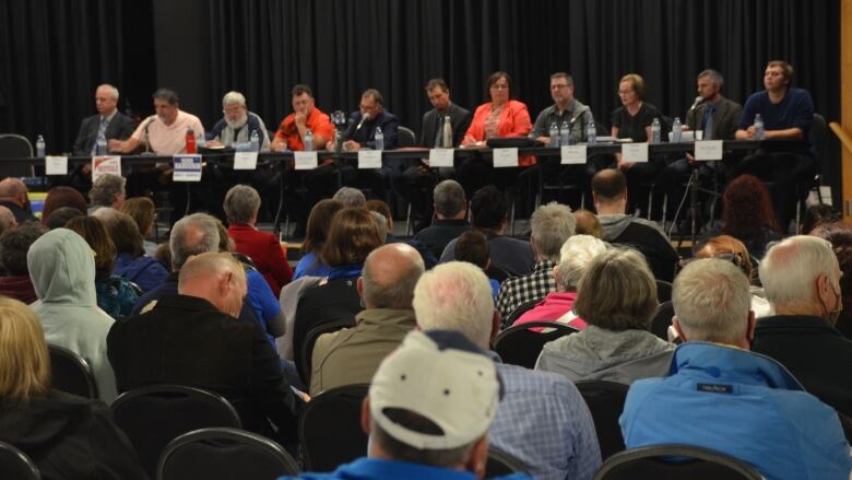 A crowd of people sit in the arena hall in Sturgeon Falls, listening to ten mayoral and council candidates on stage. 