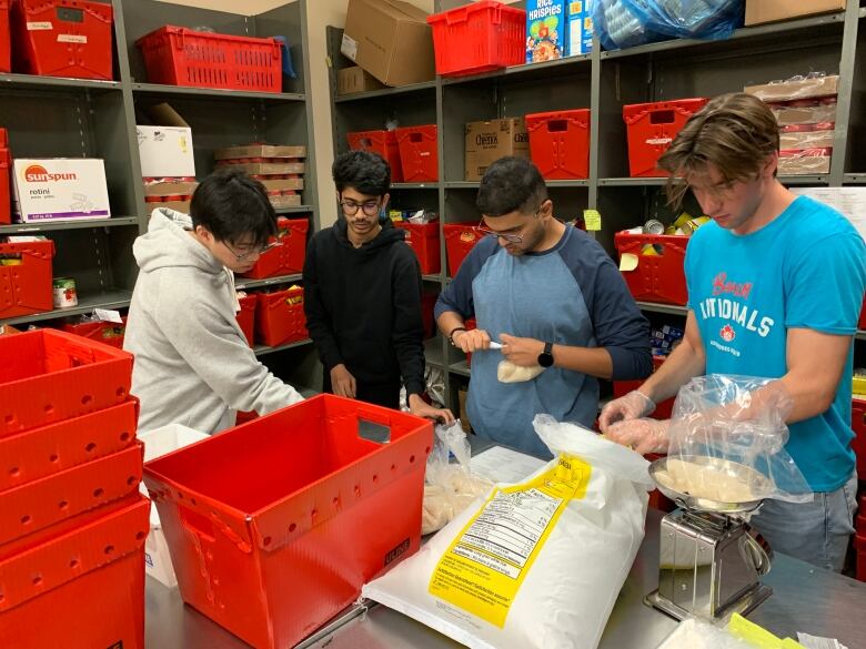 Four men stand in front of shelves. There are red bins on the table in front of them and an open bag of food. They are weighing food on a scale.