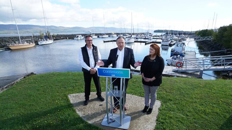 A man in a suit stands at a podium at a marina with a woman on his left and a man on his right.