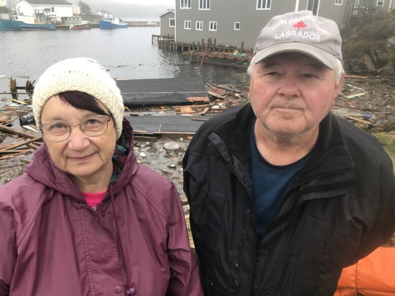 A couple stand in front of the debris left by post-tropical storm Fiona. A roof sits on the rocks of a beach, and splintered wood litters the ground.