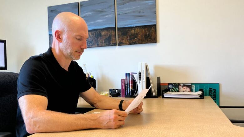 A man wearing a black short-sleeved shirt, reading from a piece of paper, while seated at a desk in an office.