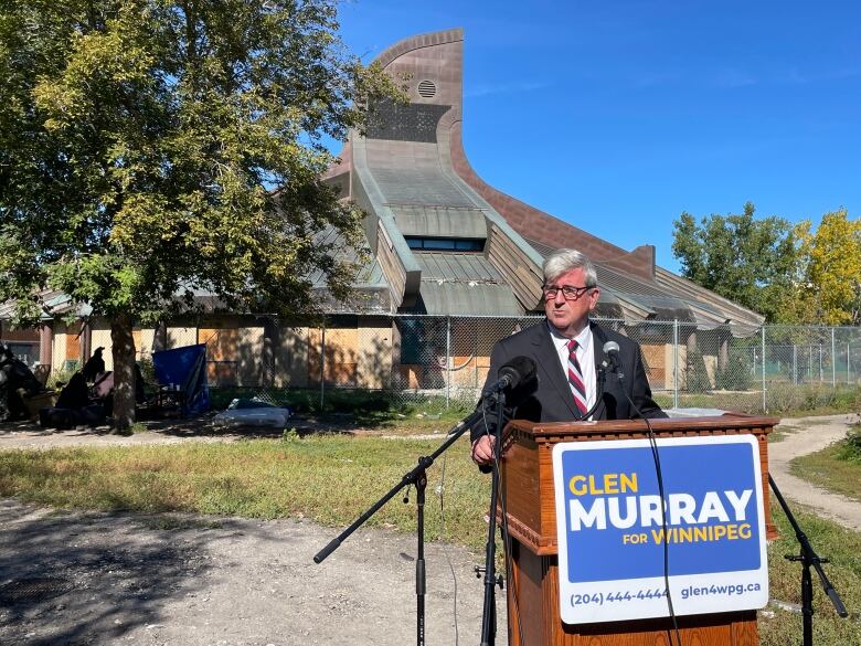 A man wearing a suit stands at a podium. Behind him, a large building with an ornate roof is surrounded by a fence.