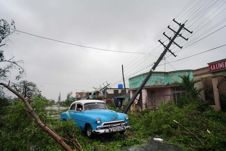 A bright blue car sits among storm debris, with fallen power lines to the right. 