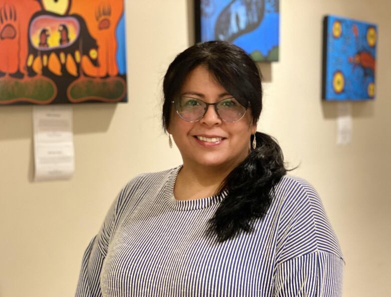 A woman with dark hair and glasses stands in front of a wall of Indigenous art.