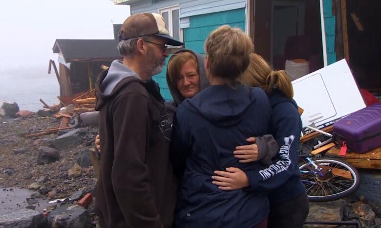 Four people in a group hug stand in front of the ruins of a home. The ocean can be seen in the top left.