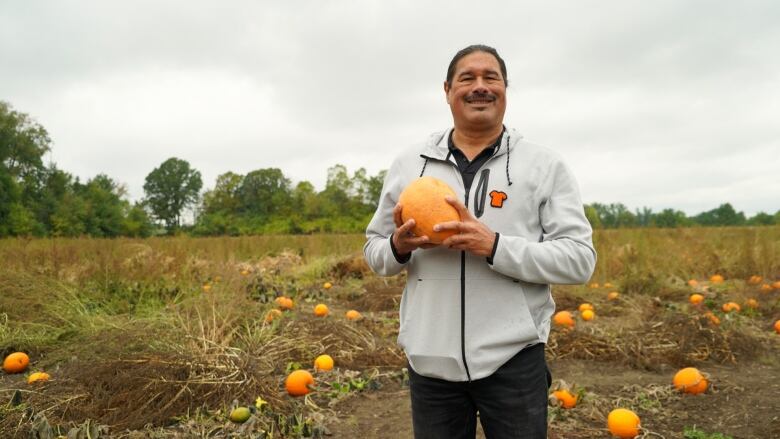 An man stands in a field of pumpkins holding a squash on a cloudy day.