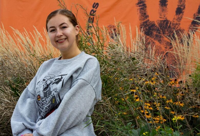 A young woman in a sweatshirt smiles at the camera. Behind her an orange sign with a child's handprint.