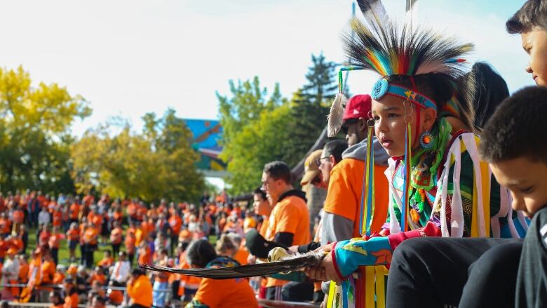 A boy in regalia looks on as hundreds of people wearing orange shirts are seen in the background.