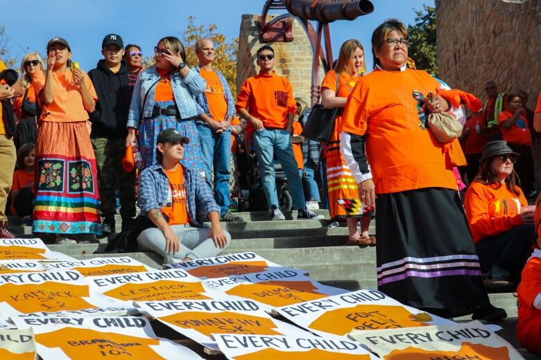 People wearing on orange shirts stand on steps, while leaving space for a number of 'Every Child Matters' posters.