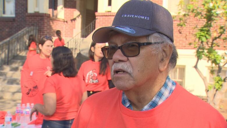 An old Indigenous man is seen wearing a hat and an orange sweater.