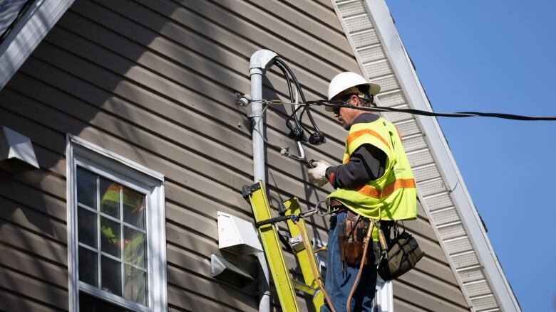 A person wears a safety vest and hard hat while on a ladder that's leaning on a house.