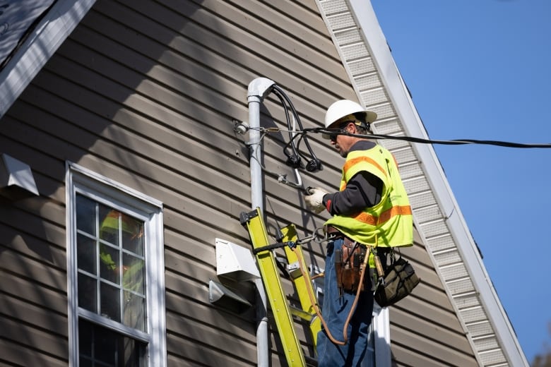 A person wears a safety vest and hard hat while on a ladder that's leaning on a house.