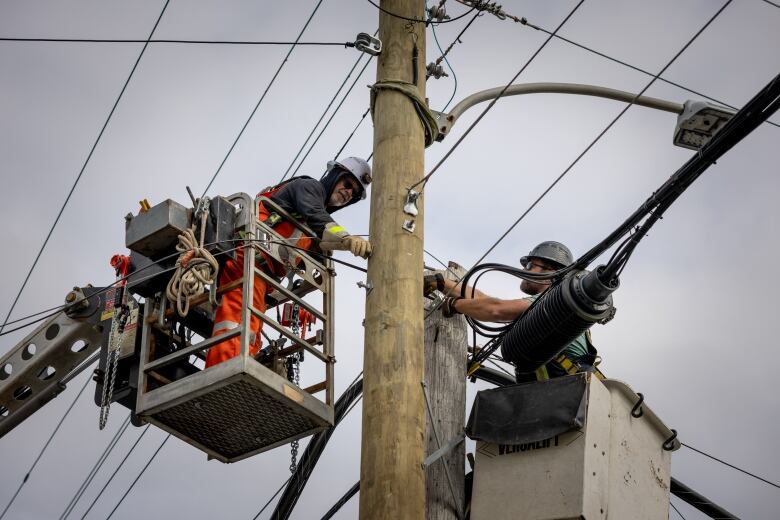 Two men wearing hard hats are seen on either side of a power pole. Both are in box lifts and have their arms outstretched toward the pole wile they hold tools.