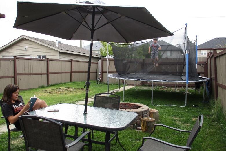 A young boy jumps on a trampoline while a young woman reads a book. 