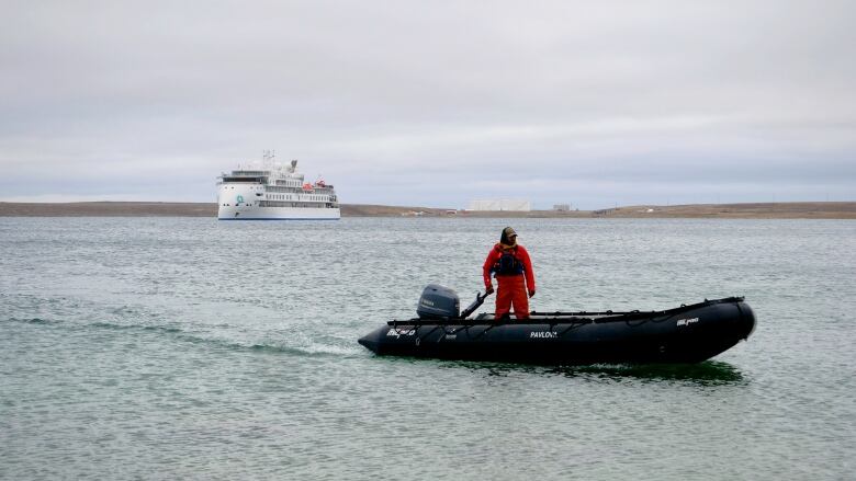 A man in a small motorized boat steers toward shore while a large cruise ship sits anchored distantly behind him.