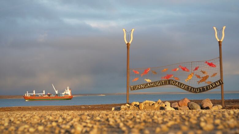 A rocky beach with a large ship distantly in the water and a sign on the shores written in Inuktitut. 