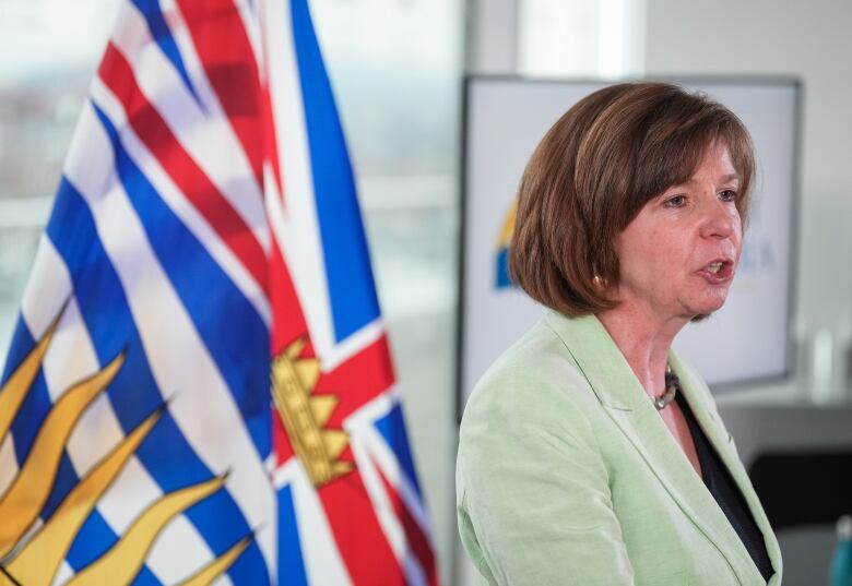 A woman in a green suit with a B.C. flag behind her speaks to reporters who are off camera.