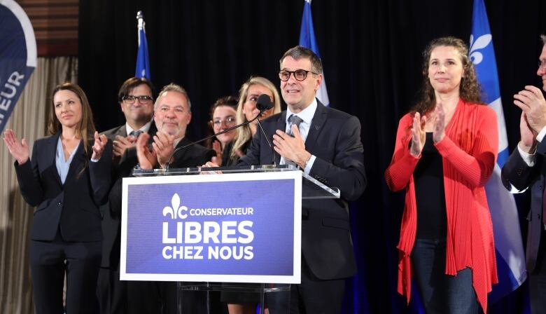 A group of people standing on a stage. A man in the centre is speaking on a podium.