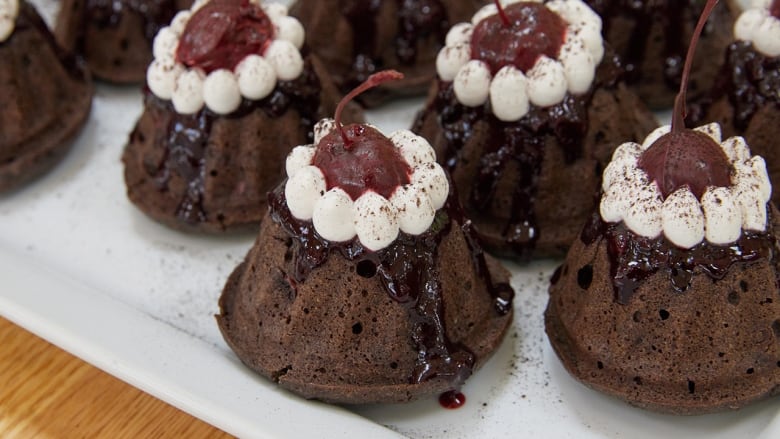 Mini chocolate bundt cakes topped with cherry sauce, whipped cream and a chocolate-covered cherry sitting on a white dessert platter.