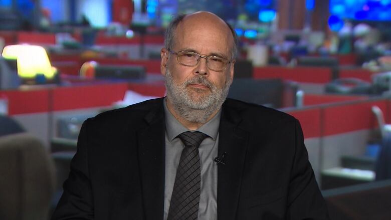 A balding man with a beard is seated with a blurred image of a newsroom behind him.