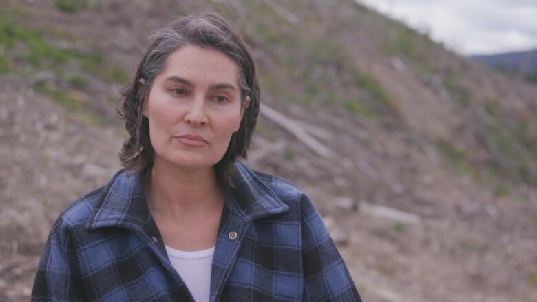 A woman with a pensive face looks off-camera with a logged-out hill in the background.