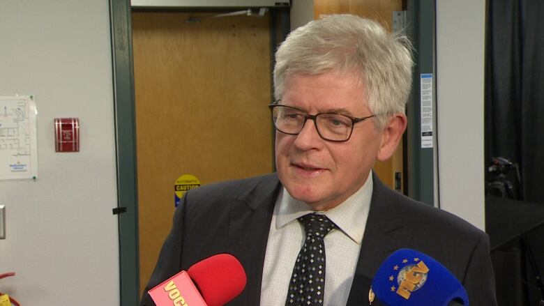 A grey-haired man stands in front of several microphones as part of a media scrum.