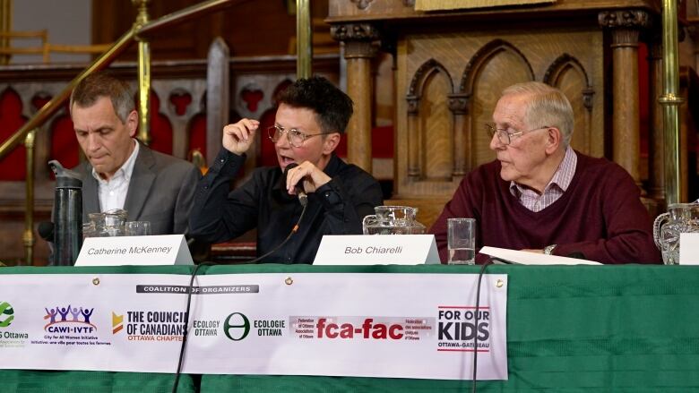 Three politicians sit at a table. The middle one is speaking on a microphone.