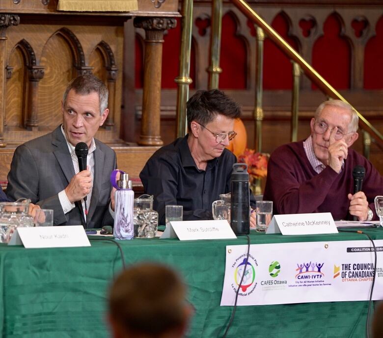 Three politicians sit at a table. The left one is speaking on a microphone.