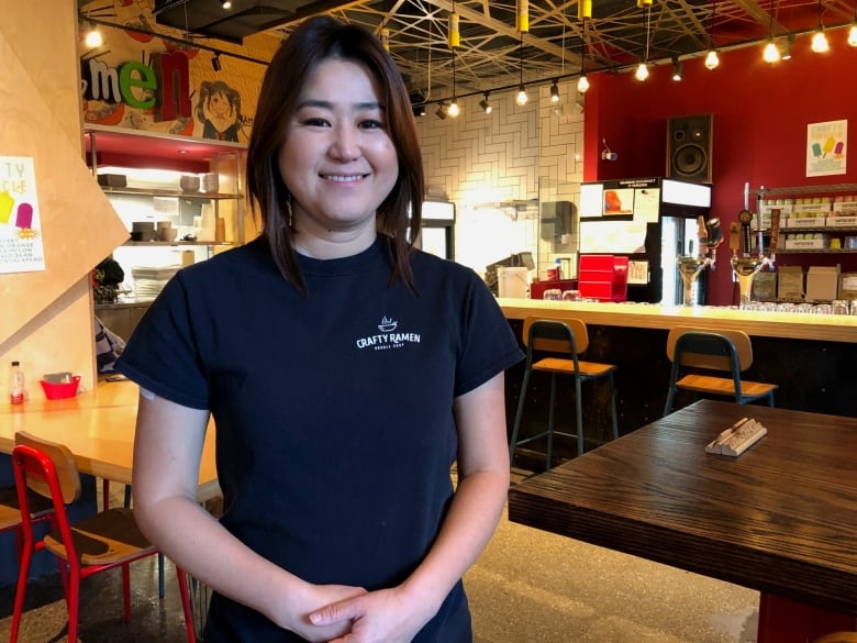 A woman wearing a black T-shirt stands in the center of a Ramen shop.