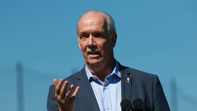 John Horgan gestures at a podium. He is a white man with a white moustache.