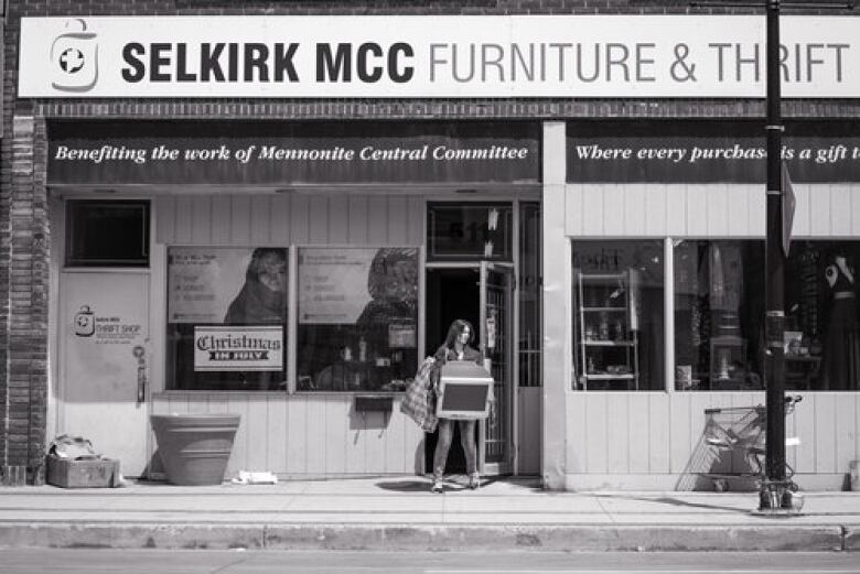 A black-and-white photo shows a wide shot of a woman walking out of a store carrying a TV. The sign on the store read 