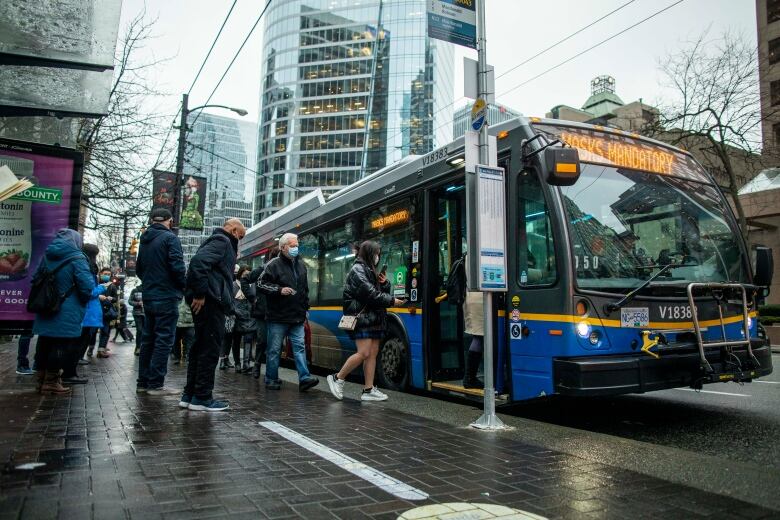 People are seen boarding a bus, wearing masks, on a rainy day.