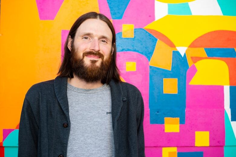 A man with long hair and a beard stands in front of a colourful wall.