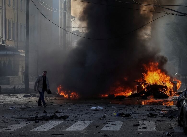 A man, shrouded in black smoke, crosses the street in front of a pile of flaming debris. 