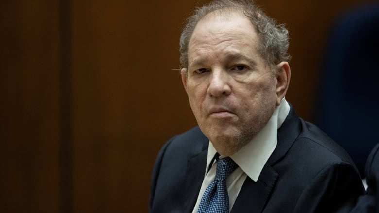 Close up of an unsmiling individual sitting at courtroom table wearing a blue suit and blue tie.