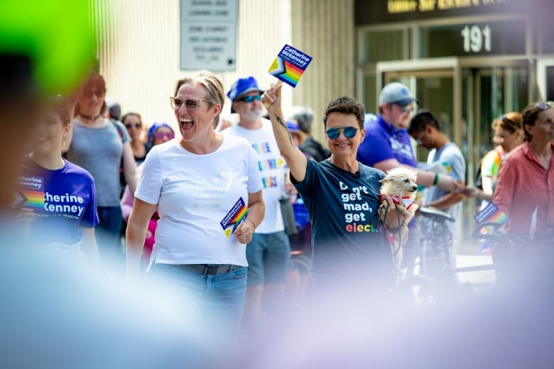 People wave flags, hold rainbow-coloured signs and smile in the middle of an outdoor celebration.