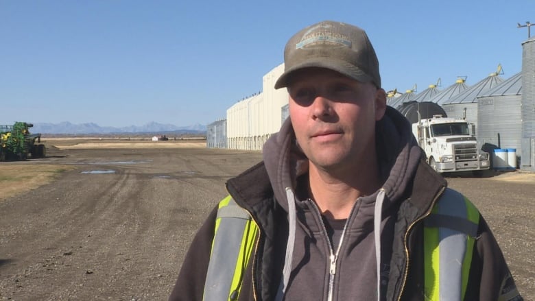 A man stands in a field with grain bins behind him.