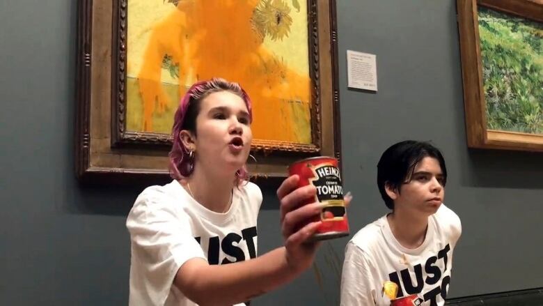 Two protesters are standing in front of Vincent Van Gogh's famous painting Sunflowers at the National Gallery in London. One of them is holding up a can of tomato soup, which was just thrown at the painting. The painting is stained by soup.