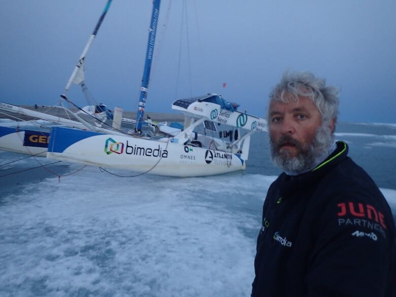 A cold-looking man looks at the camera. Behind him, a catamaran is pulled up on ice.